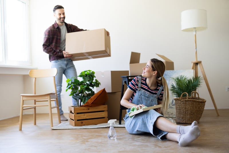 A man and a woman smile while carrying boxes into a new house.
