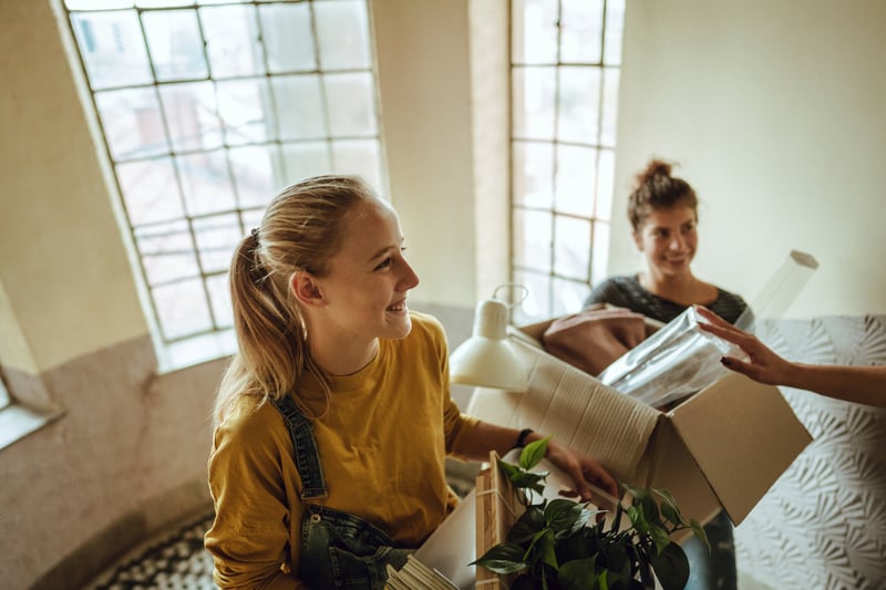 Two women smile while carrying boxes into a new house.