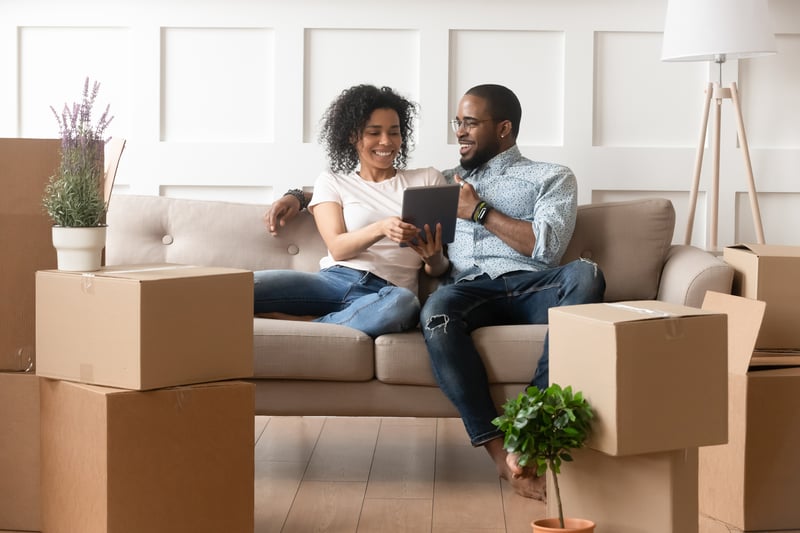 A man and a woman sit on a sofa surrounded by moving boxes.