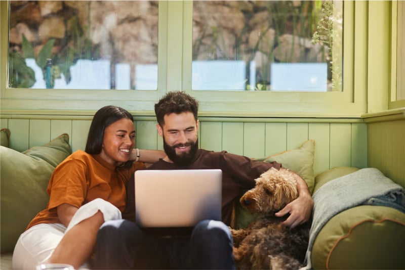 A woman, man, and a dog sit on a couch and look at a laptop. 