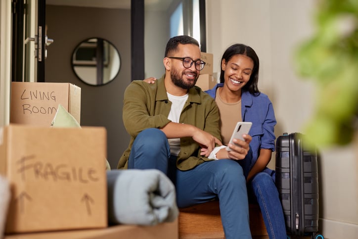 A man and a woman smile at a phone while moving into a house.