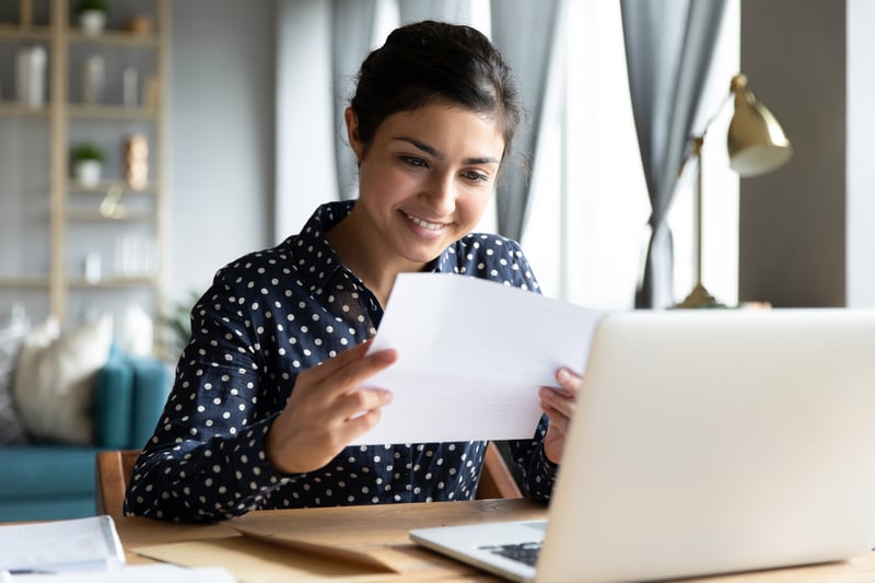 A young woman smiling at a paper while at a laptop.