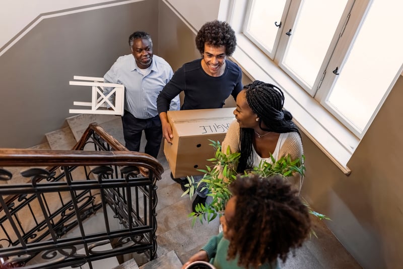 Three people carry boxes, furniture, and a plant up a stairwell.