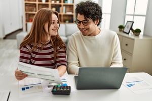 A man and a woman sit at a desk and smile while using a laptop and calculator.