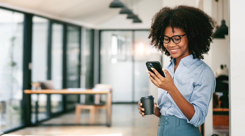 A woman smiles at a cell phone while holding a coffee mug.