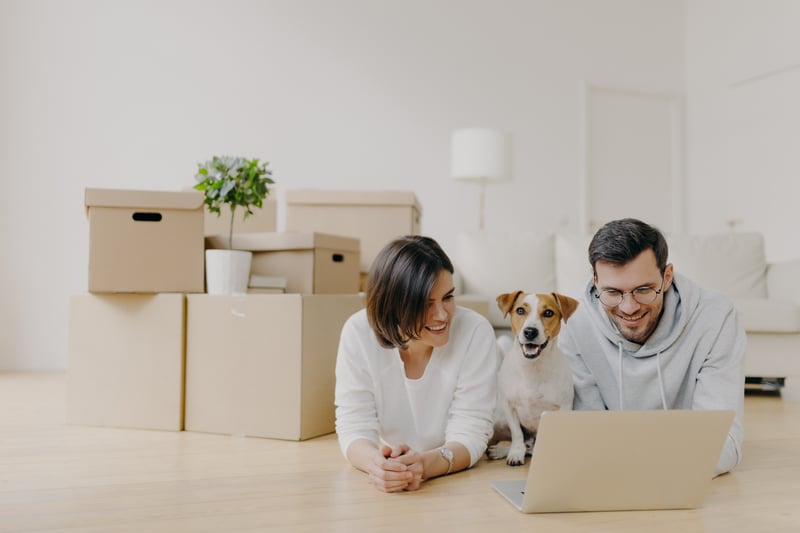 A woman and man smile at a laptop and while on the floor with a jack russell terrior.