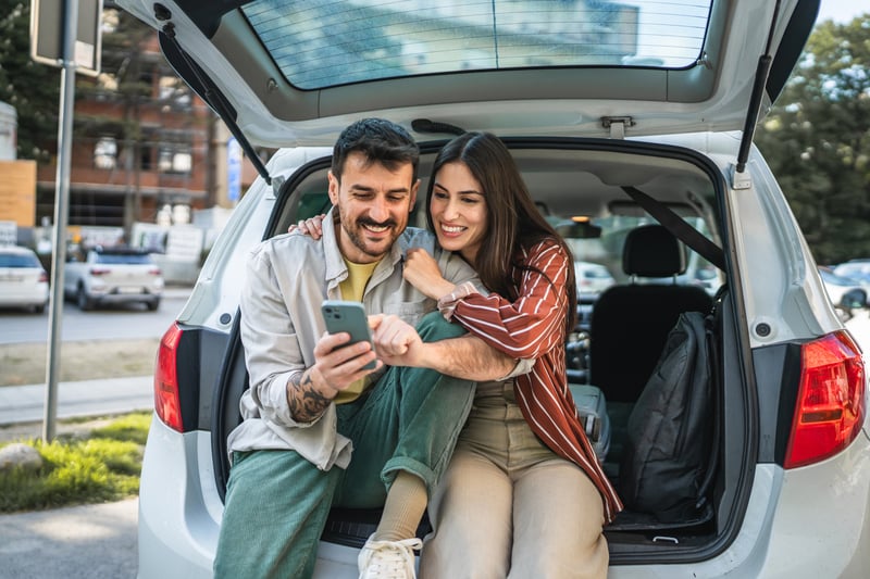 A couple sits on the rear bumper of an SUV and smiles at a phone.