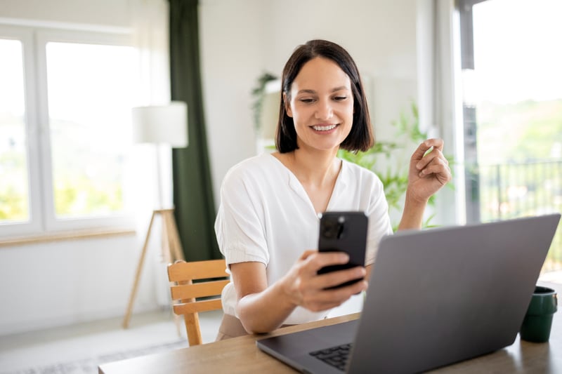 A woman smiles at a phone and laptop.
