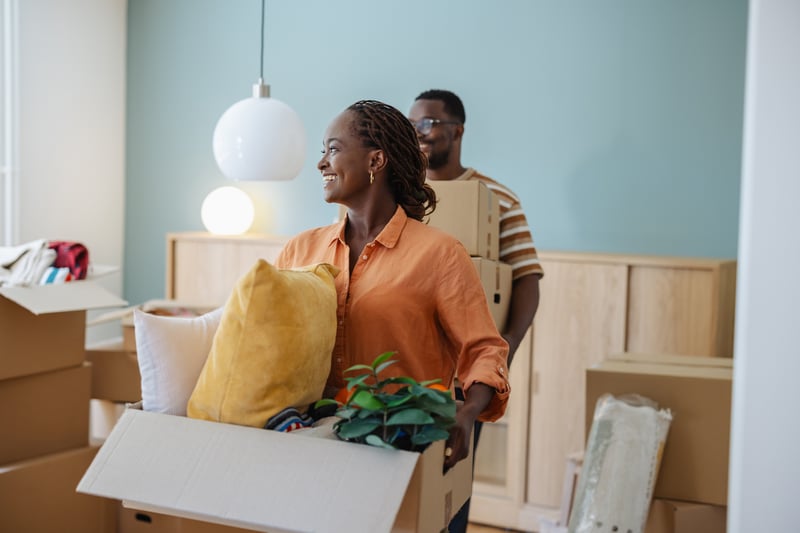 A man and a woman smile while carrying boxes into a house.