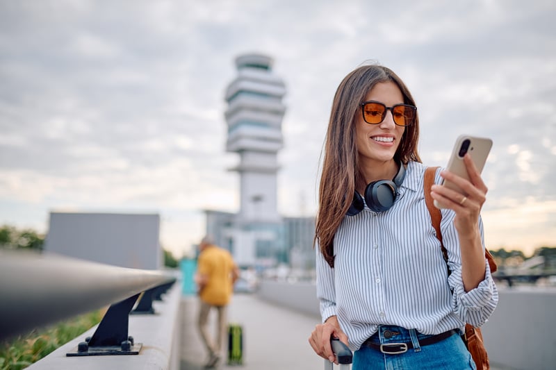 A woman at an airport smiles at a phone while looking at California refinance rates.