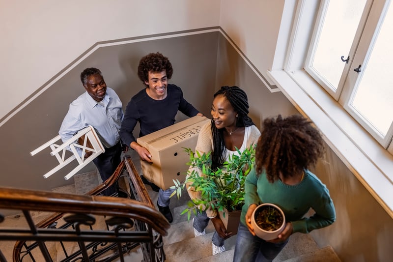 Four people carry boxes and plants up stairs into a new house.