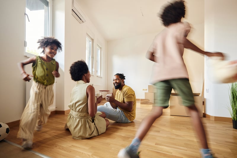 A man and a woman sit on the floor of their new living room while their kids run around.