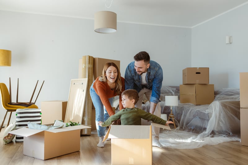 A man and a woman push a young boy in a box around a new home.