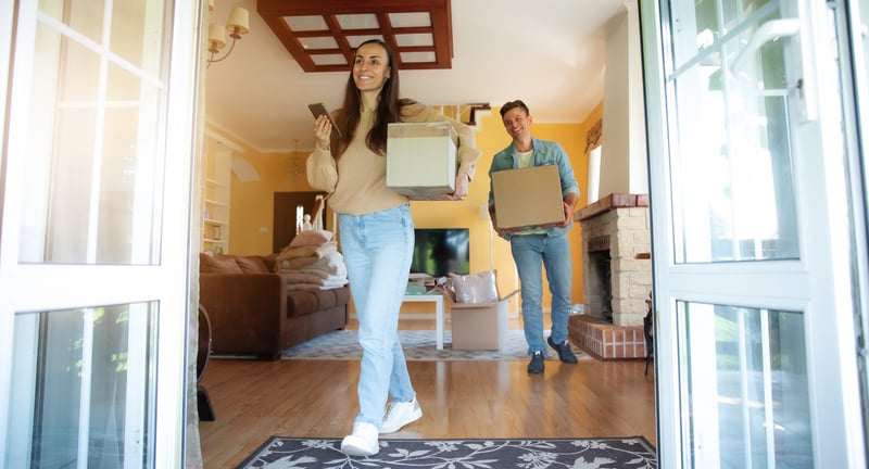 A man and a woman carry boxes into a new home.