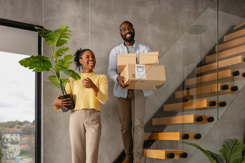 A man and a woman stand on the stairs of their new home with boxes.