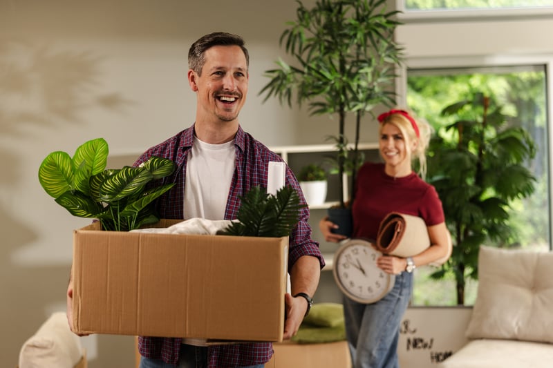 A man and a woman smile while carrying boxes into a new house.