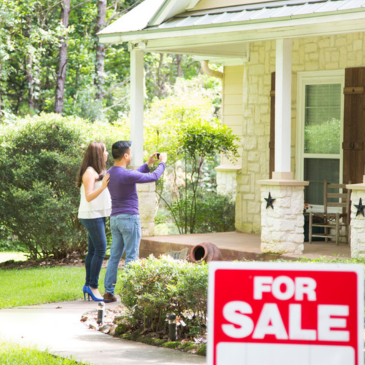 Two people stand outside a house, looking at the exterior while a red “For Sale” sign is visible in the foreground.