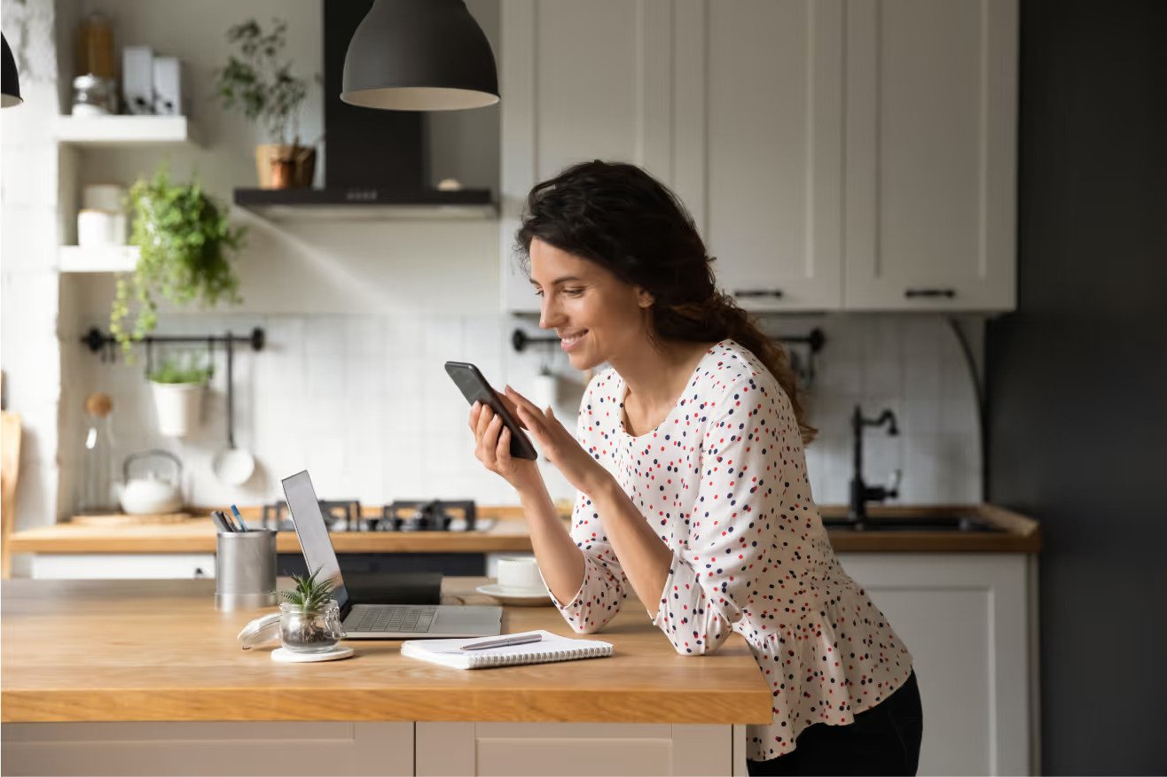 A woman with a phone and a laptop at a kitchen counter.
