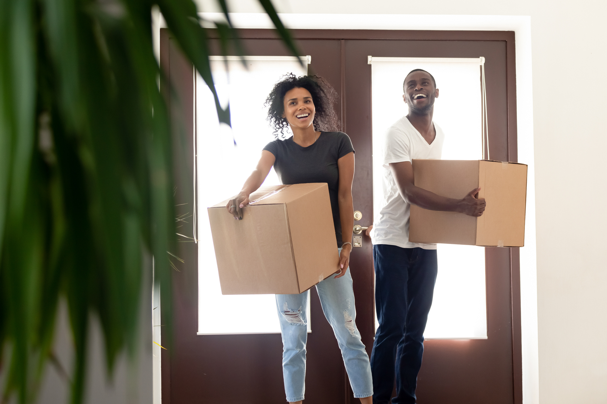 A man and a woman laugh while moving boxes into a house.