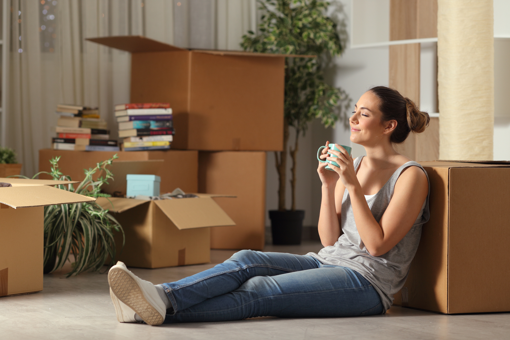 A woman smiles and drinks coffee while sitting on the floor surrounded by boxes.