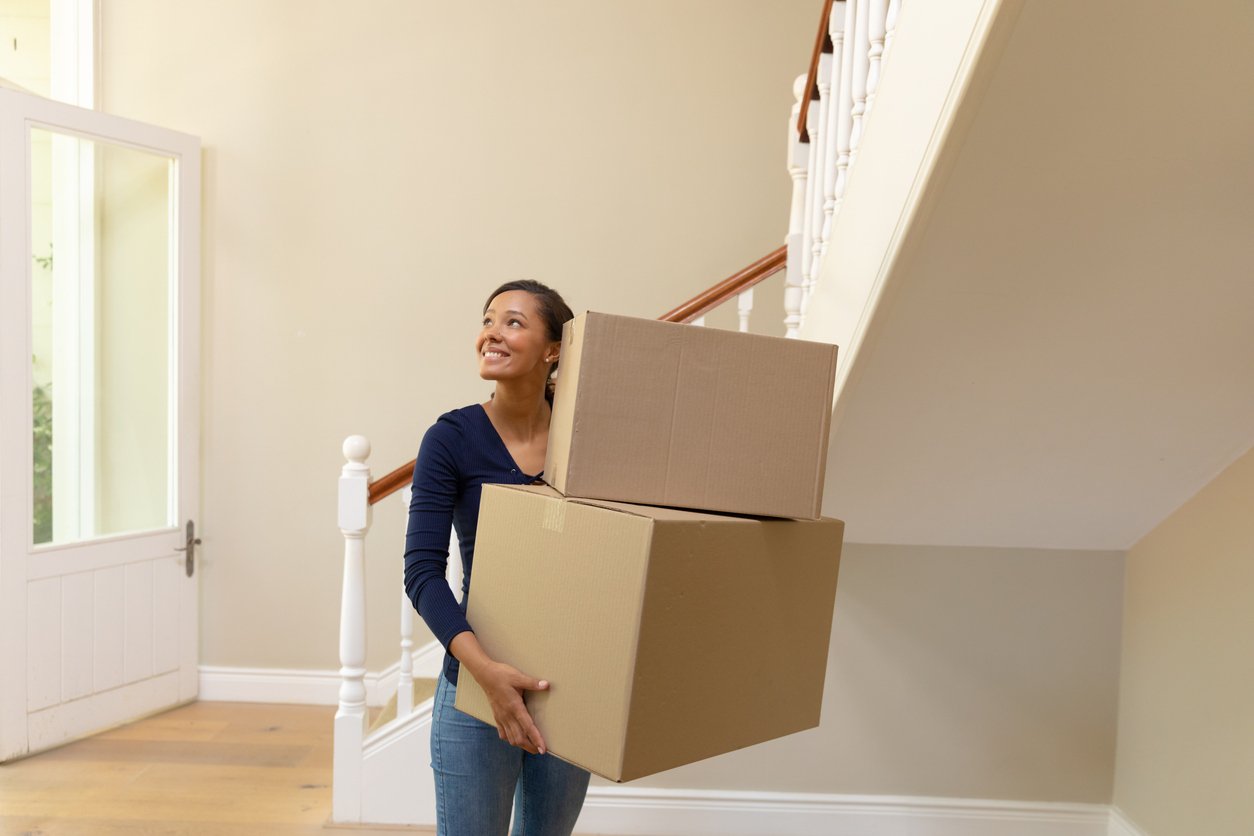 A woman smiles while carrying boxes into a house.