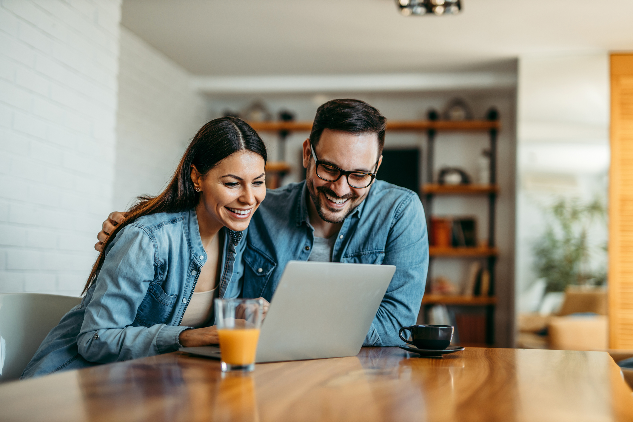 A woman and a man smile while sitting at a table and looking at a laptop.