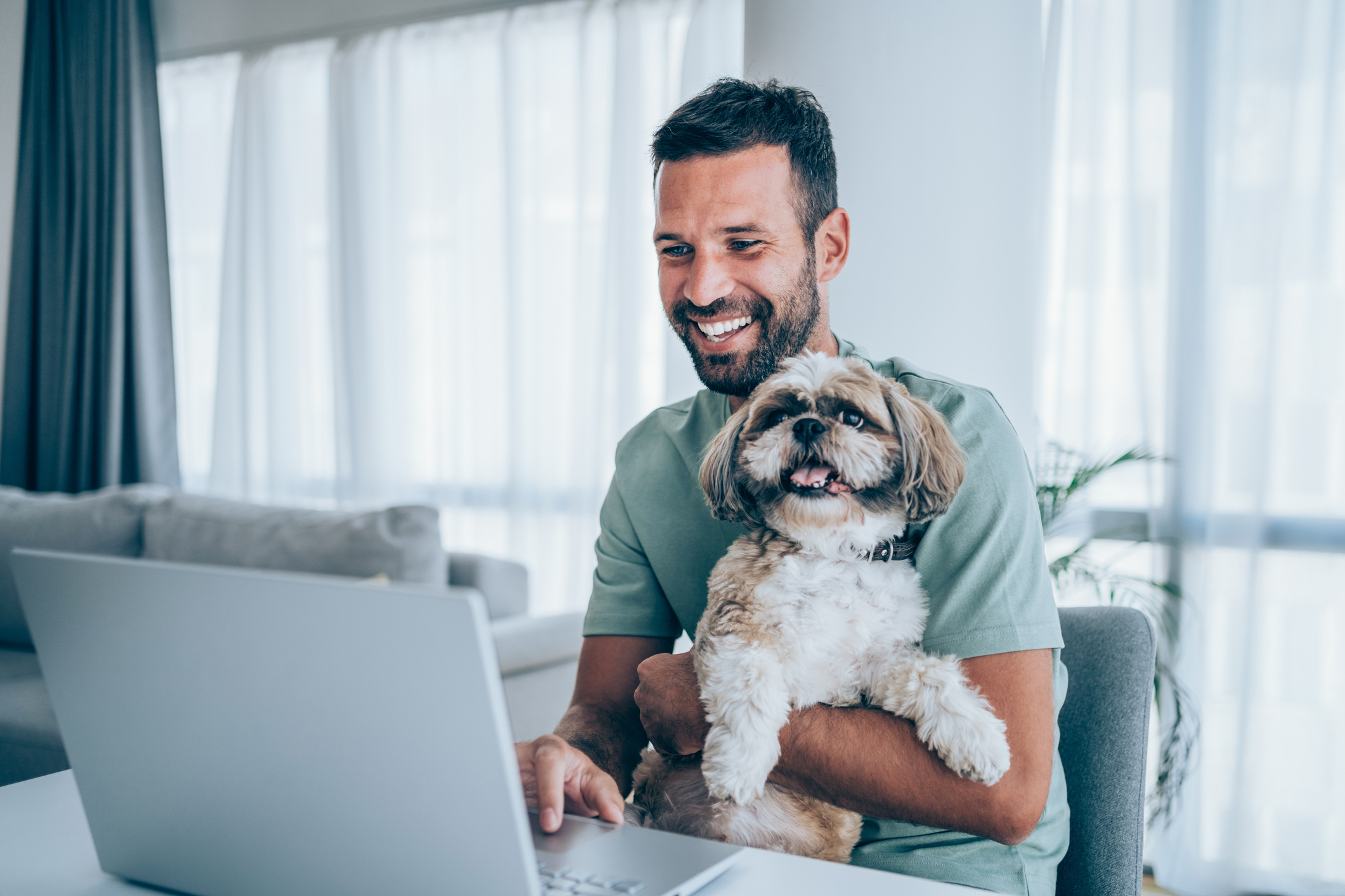 A man smiles at a laptop while holding a chunky little dog.