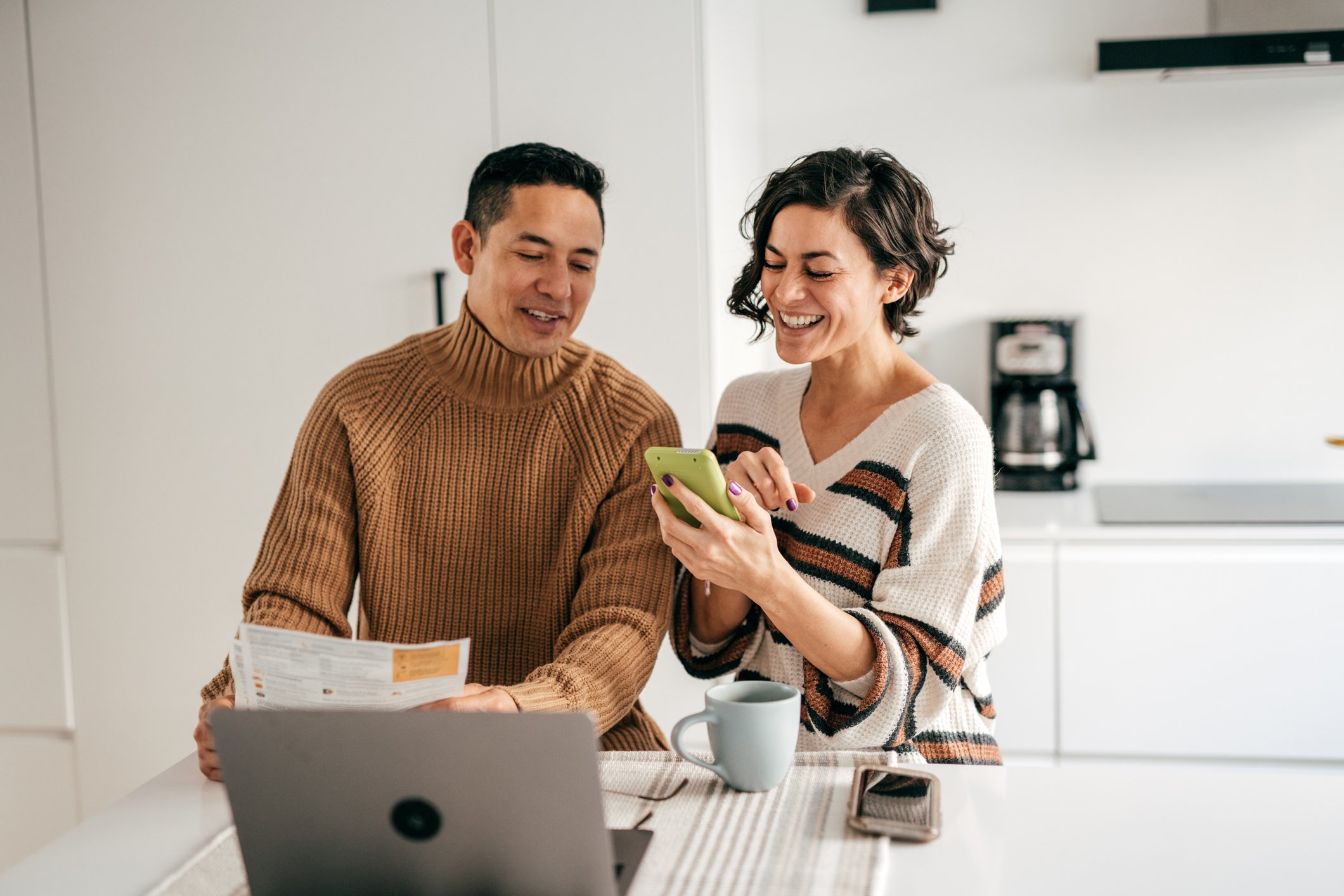 A man and a woman smiling at a cell phone while doing work at a laptop.