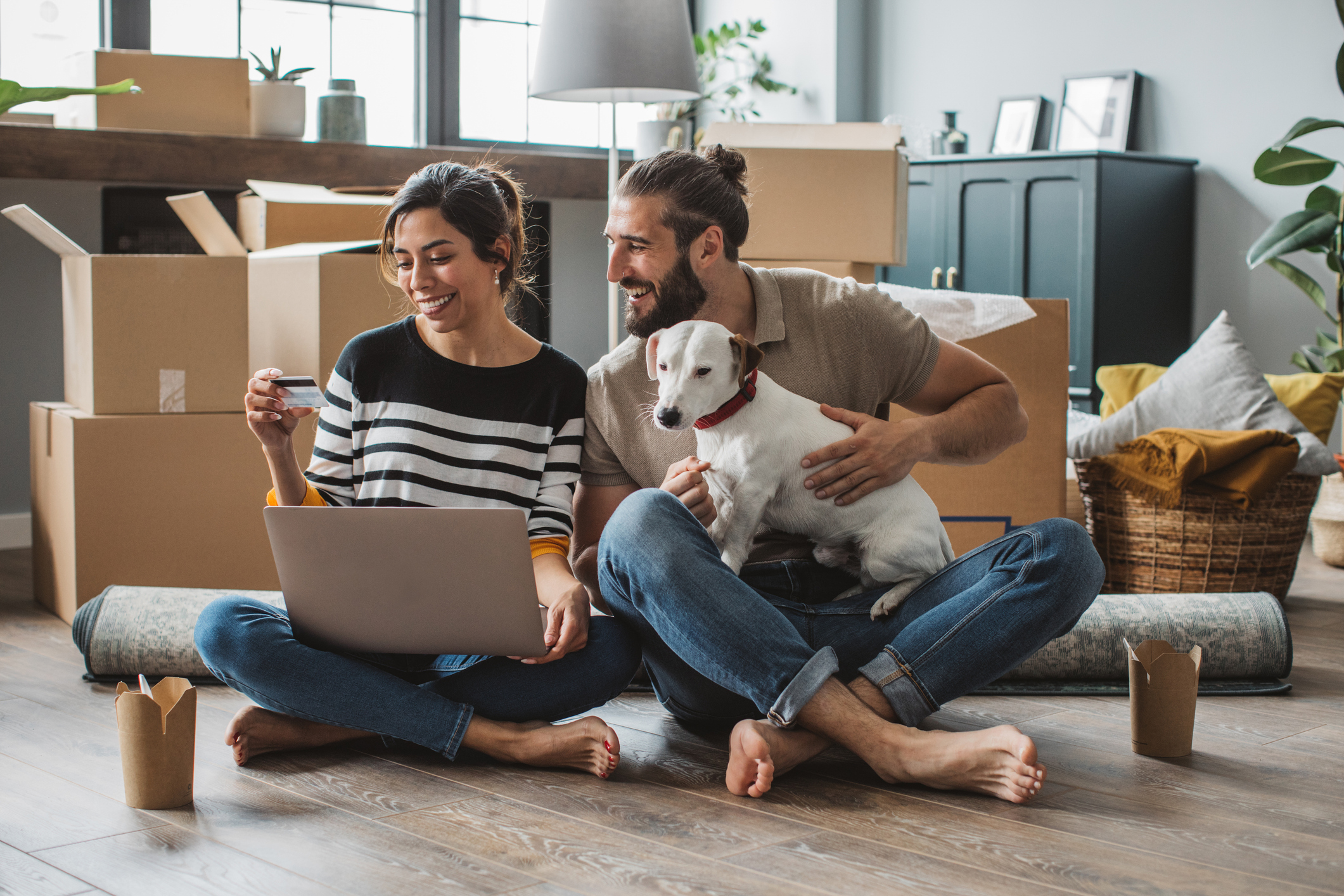 A man and a woman smile at a laptop while sitting on the floor surrounded by moving boxes with their dog.