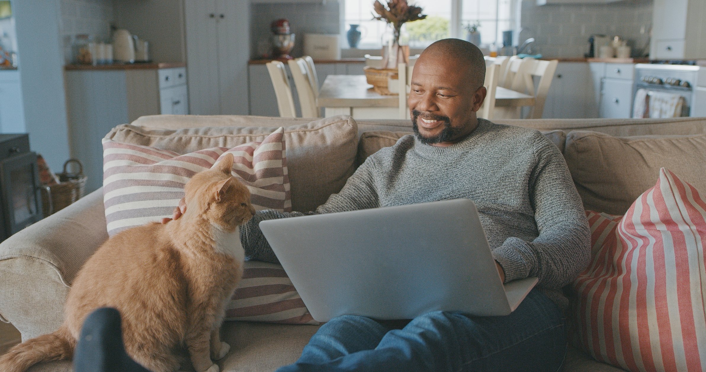 A man pets an orange cat while we works at a laptop.