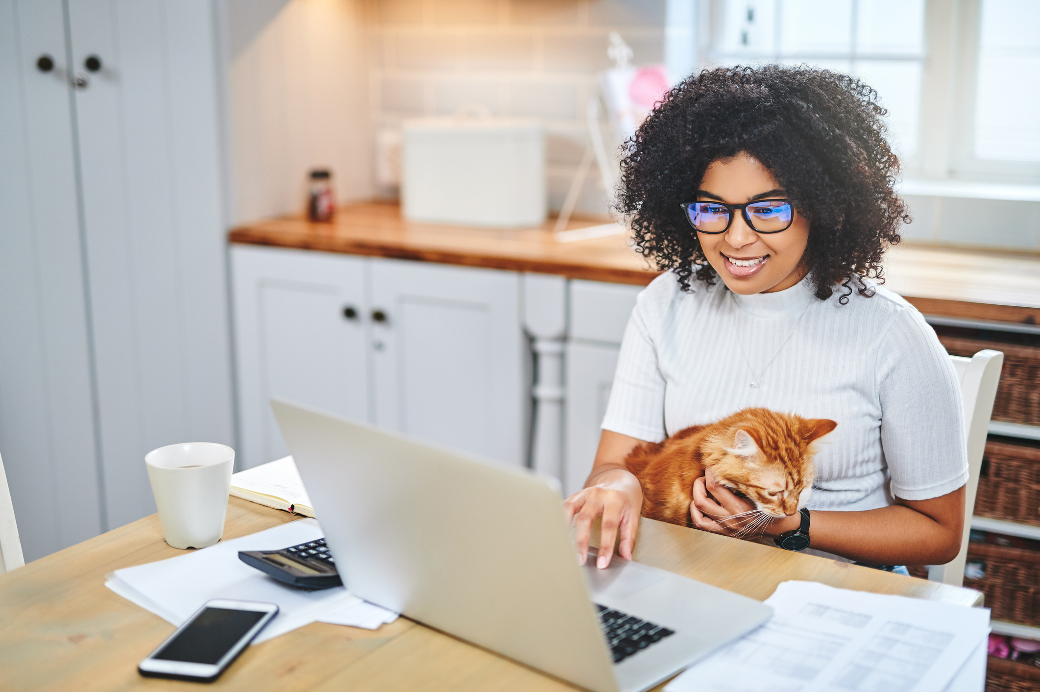 A woman smiles at a laptop while holding a sleepy orange cat.
