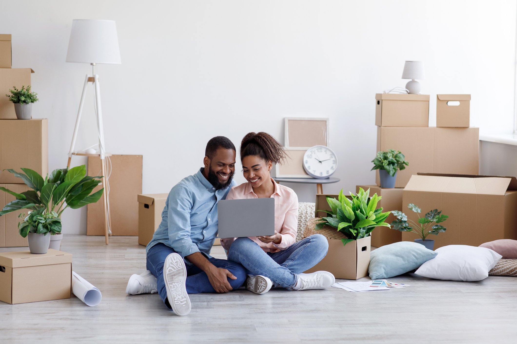 A man and a woman smile at a laptop while sitting on the floor surrounded by boxes.