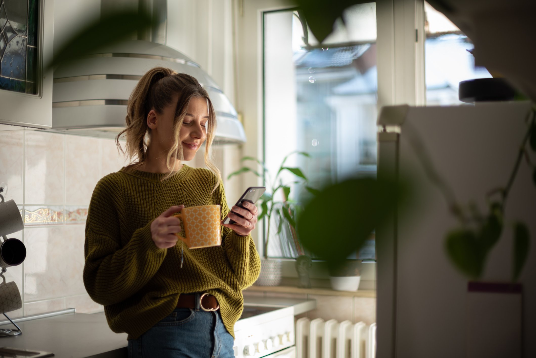 A woman smiles while looking at her phone and drinking from a mug in her kitchen.