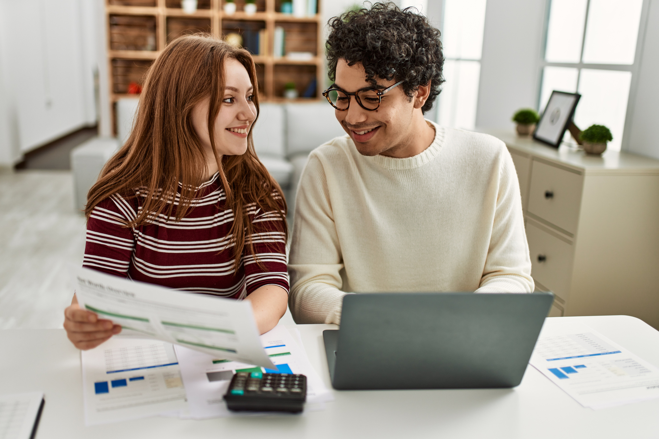 A man and a woman sit at a desk and smile while using a laptop and calculator.