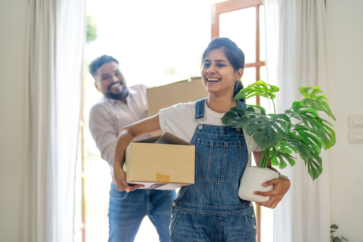 A man and a woman smile while carrying boxes and a plant into a house. 