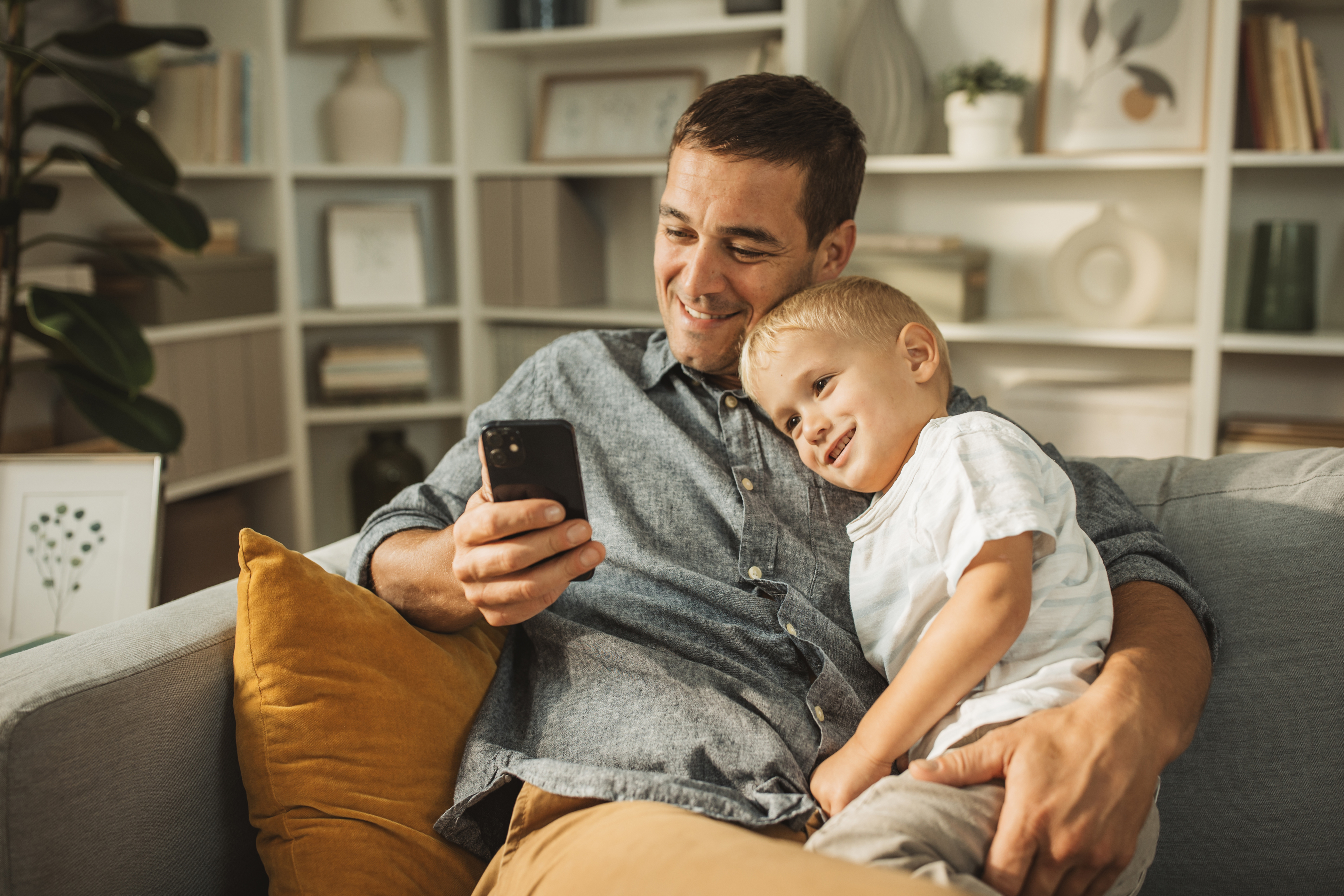 A man smiling at a phone while holding his son.