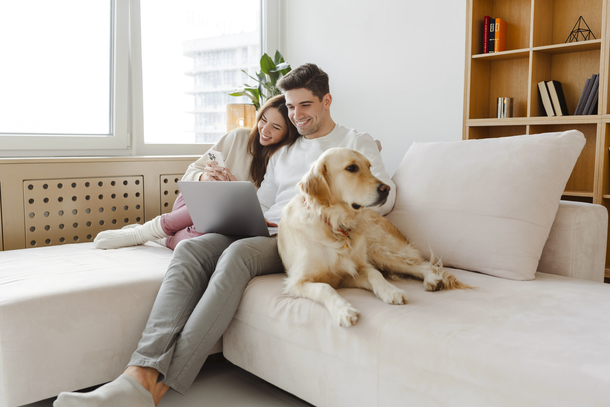 A young couple smiles at a laptop while sitting on a couch with a golden retriever.