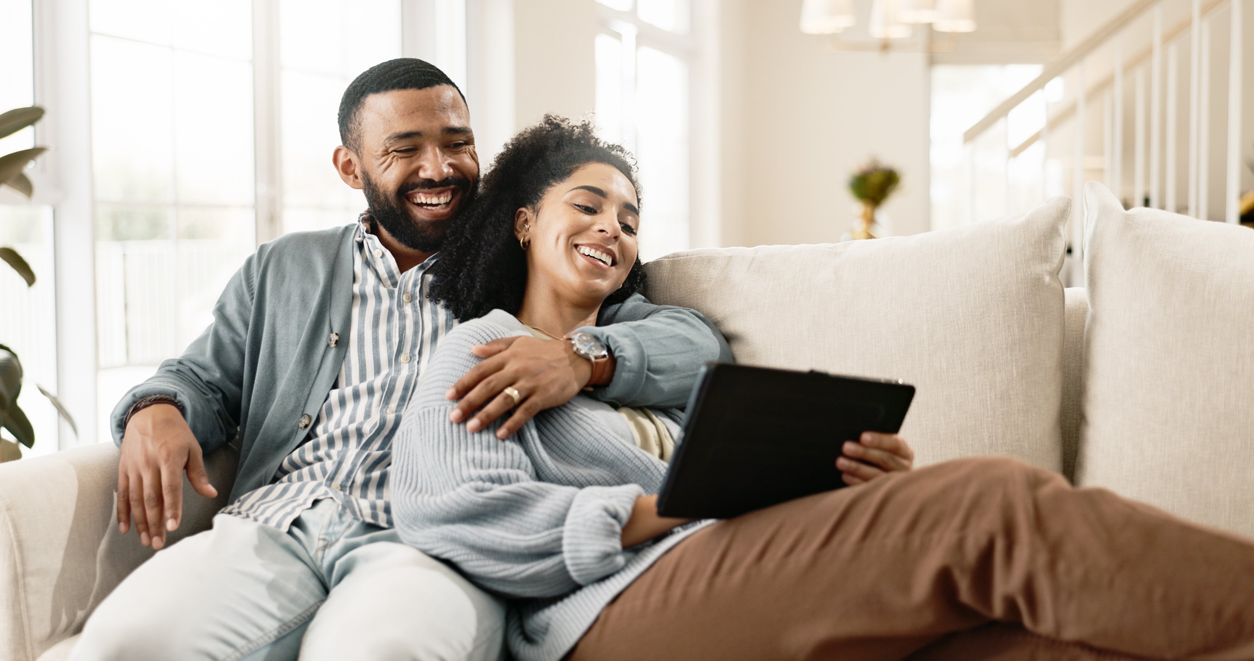 A man and woman sitting on a couch smiling at a tablet.