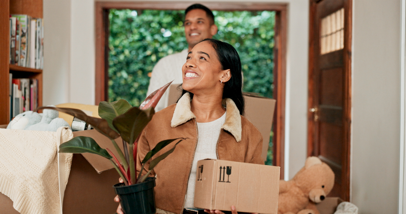 A man and a woman smile while carrying boxes into a home.