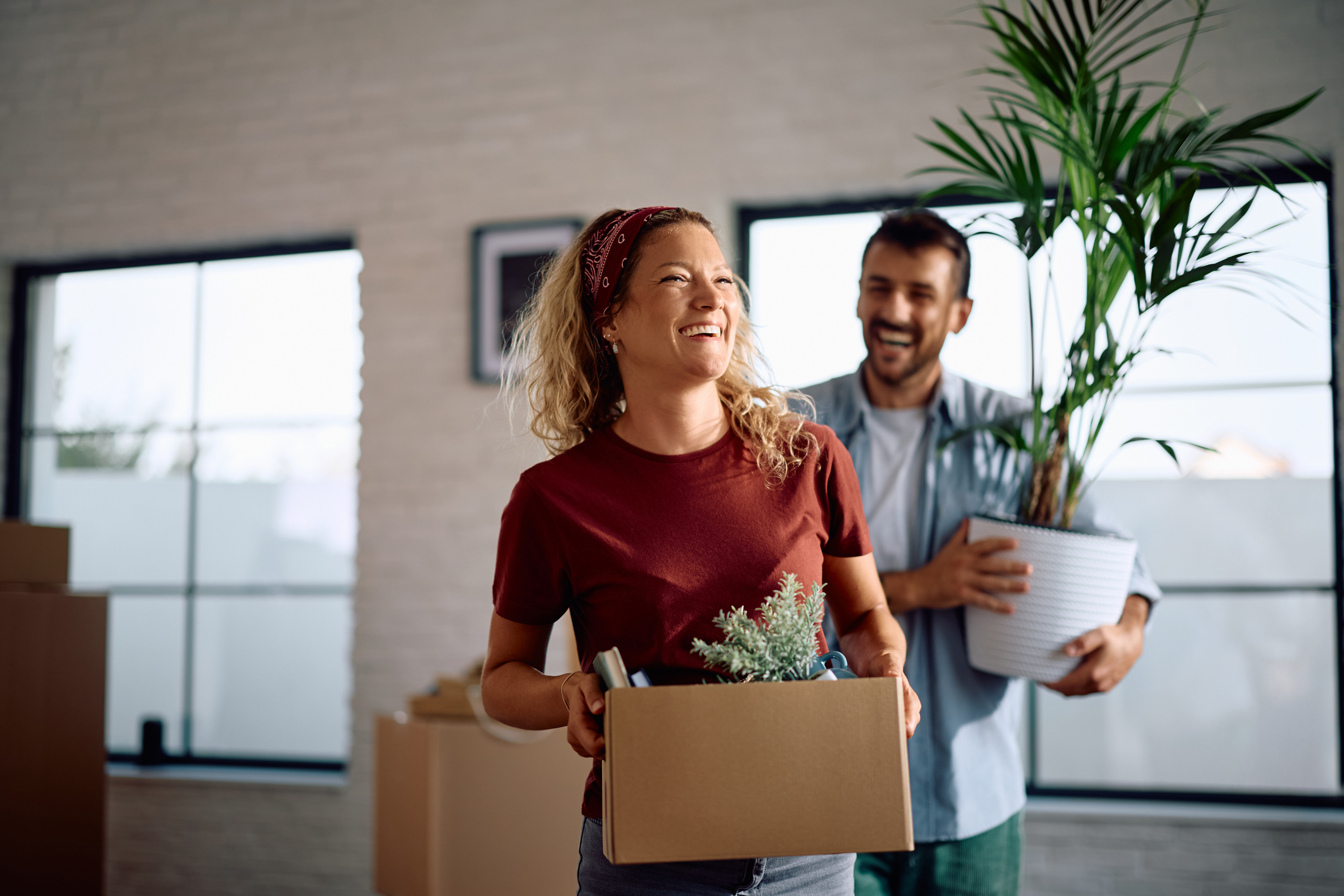 A man and a woman smiling while carrying boxes and plants into a home.