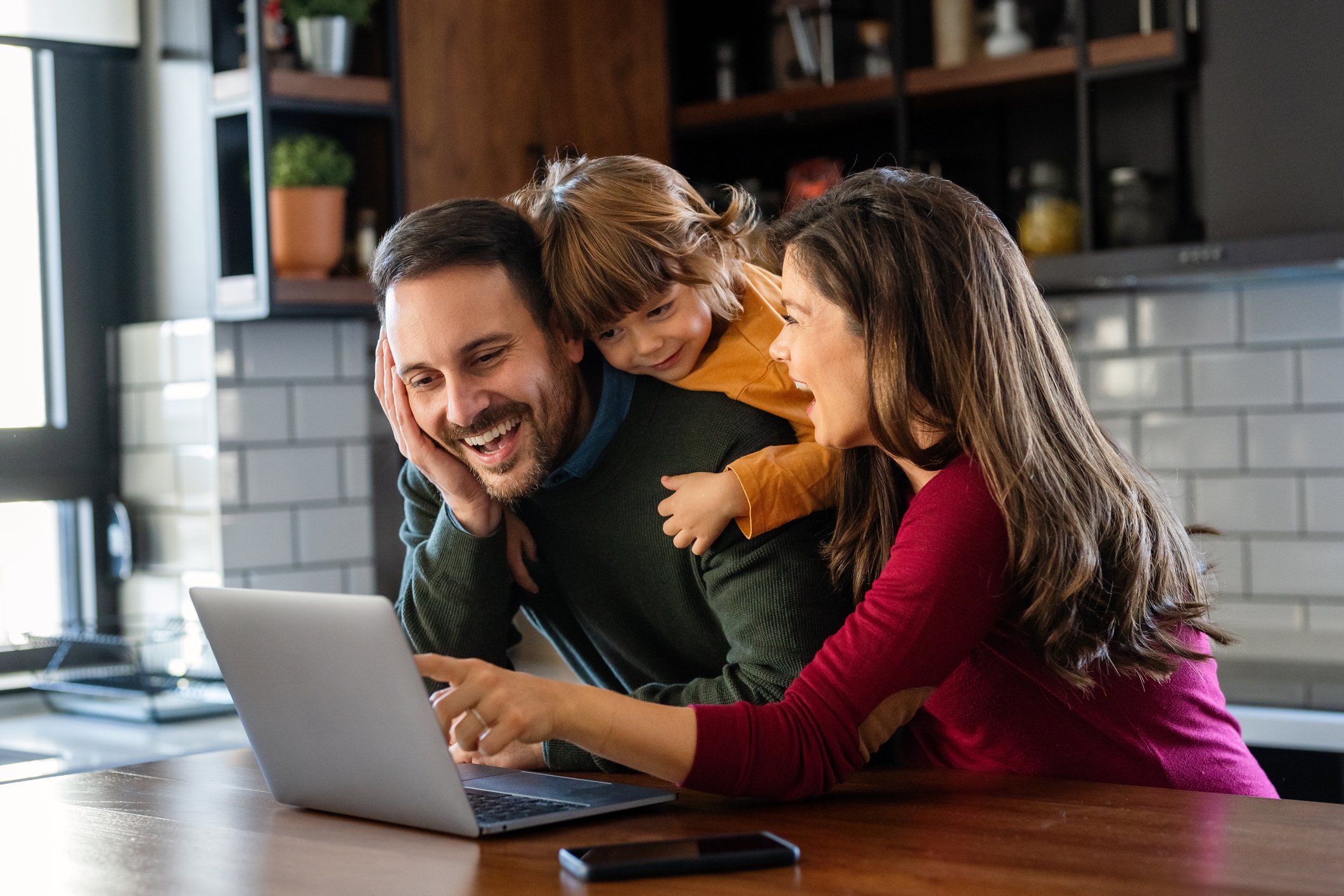 A man with a child on his back and a woman smile at a laptop.