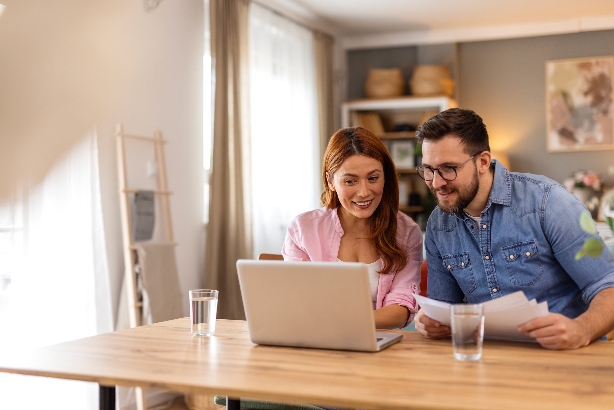 A couple smiling while looking at a laptop and holding papers.