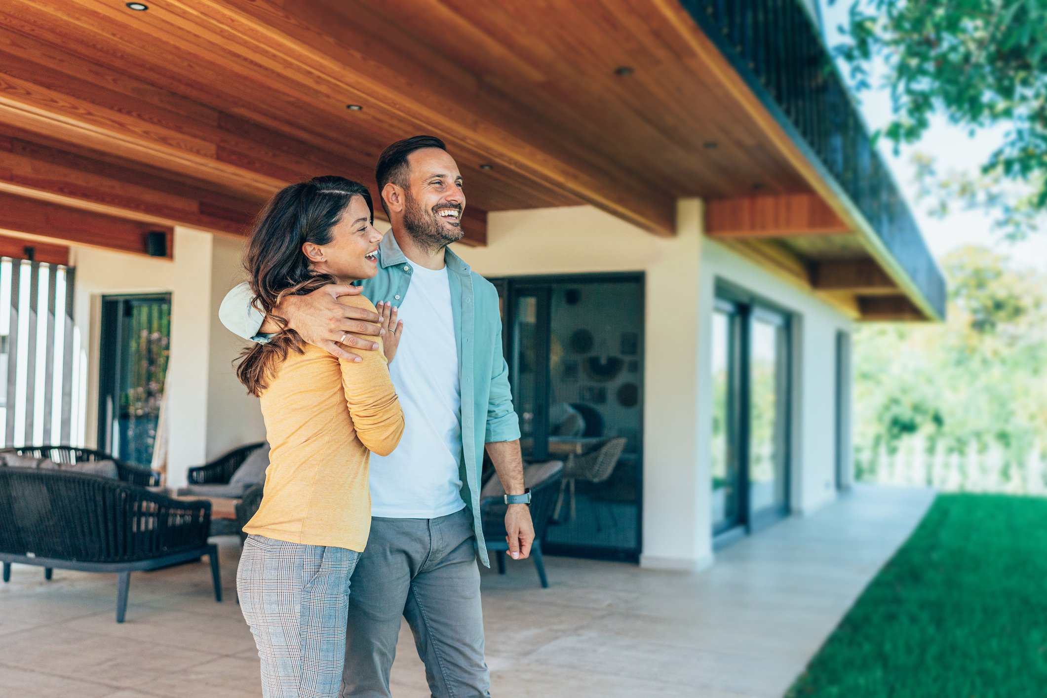A man and a woman smile with their arms around each other outside of their house. 