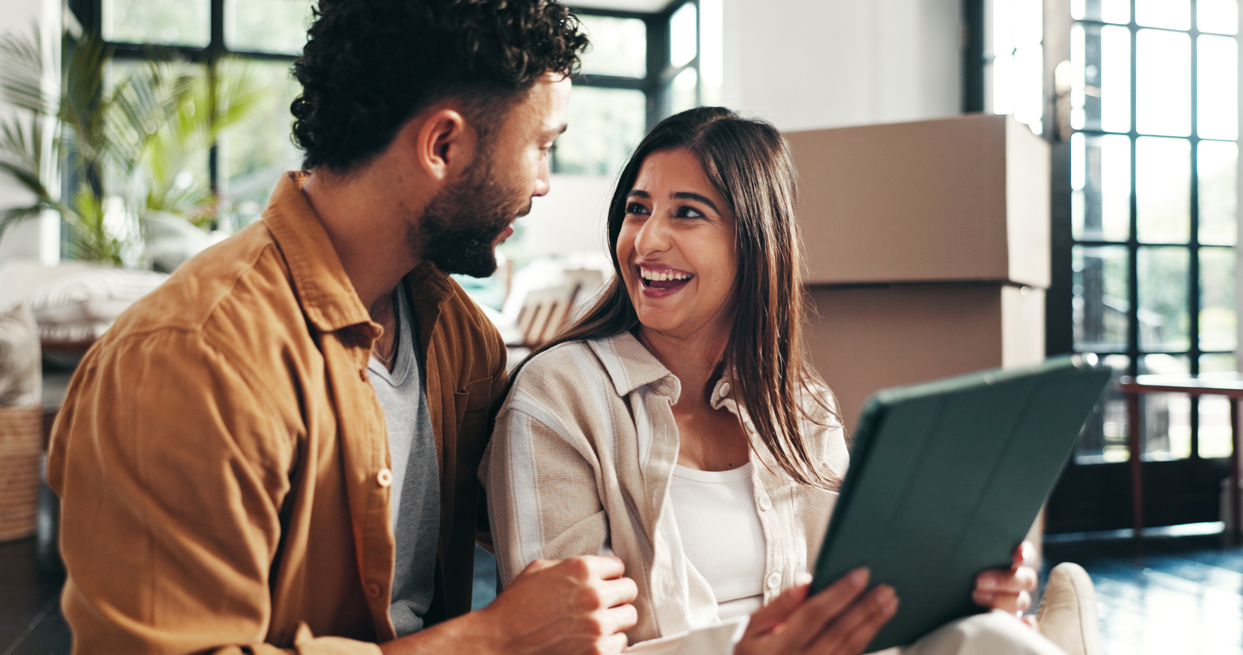 A man and a woman smile at a tablet while surrounded by moving boxes.