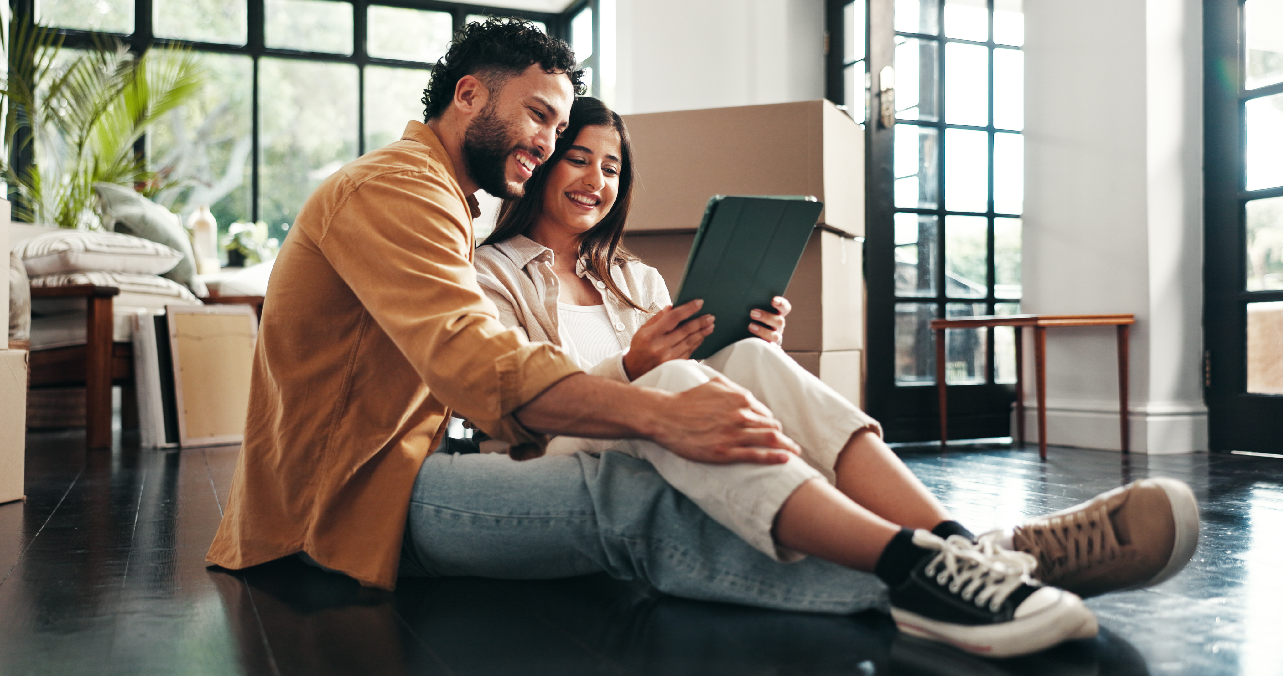 A man and a woman smile while looking at a tablet surrounded by moving boxes. 