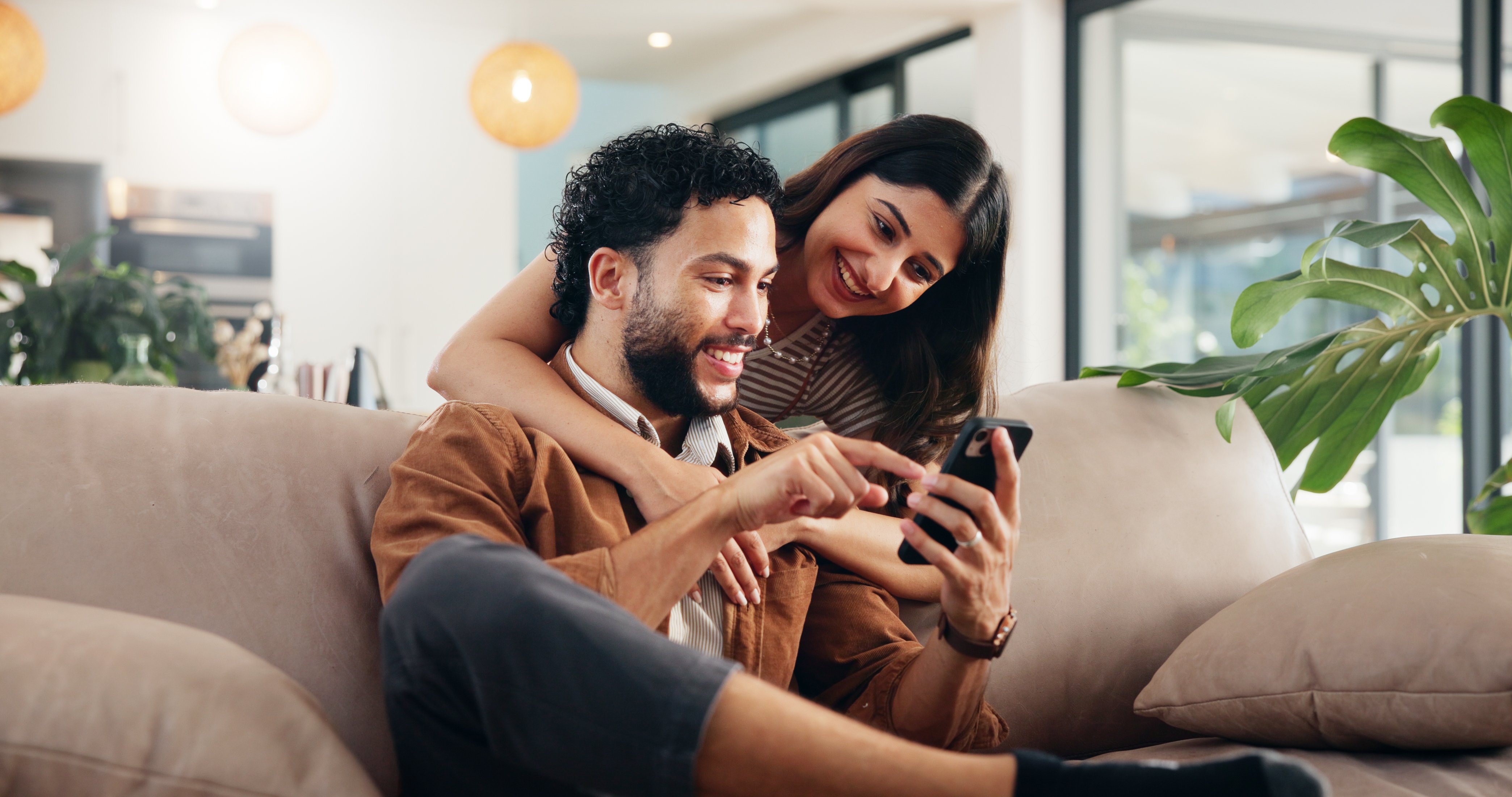 A woman puts her arms around a man while they both smile at a cell phone