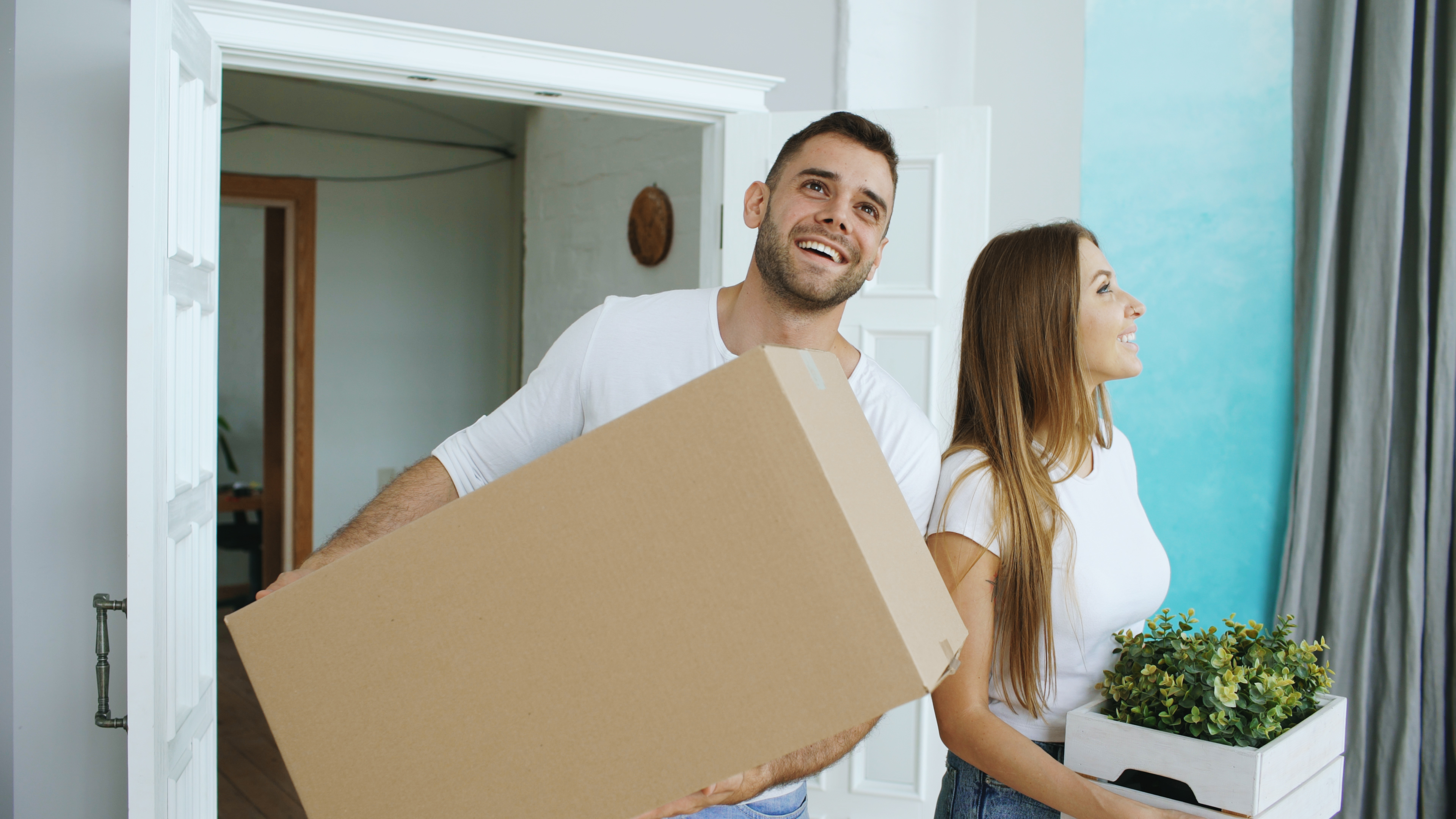 A man and woman smile while carrying boxes into a house.