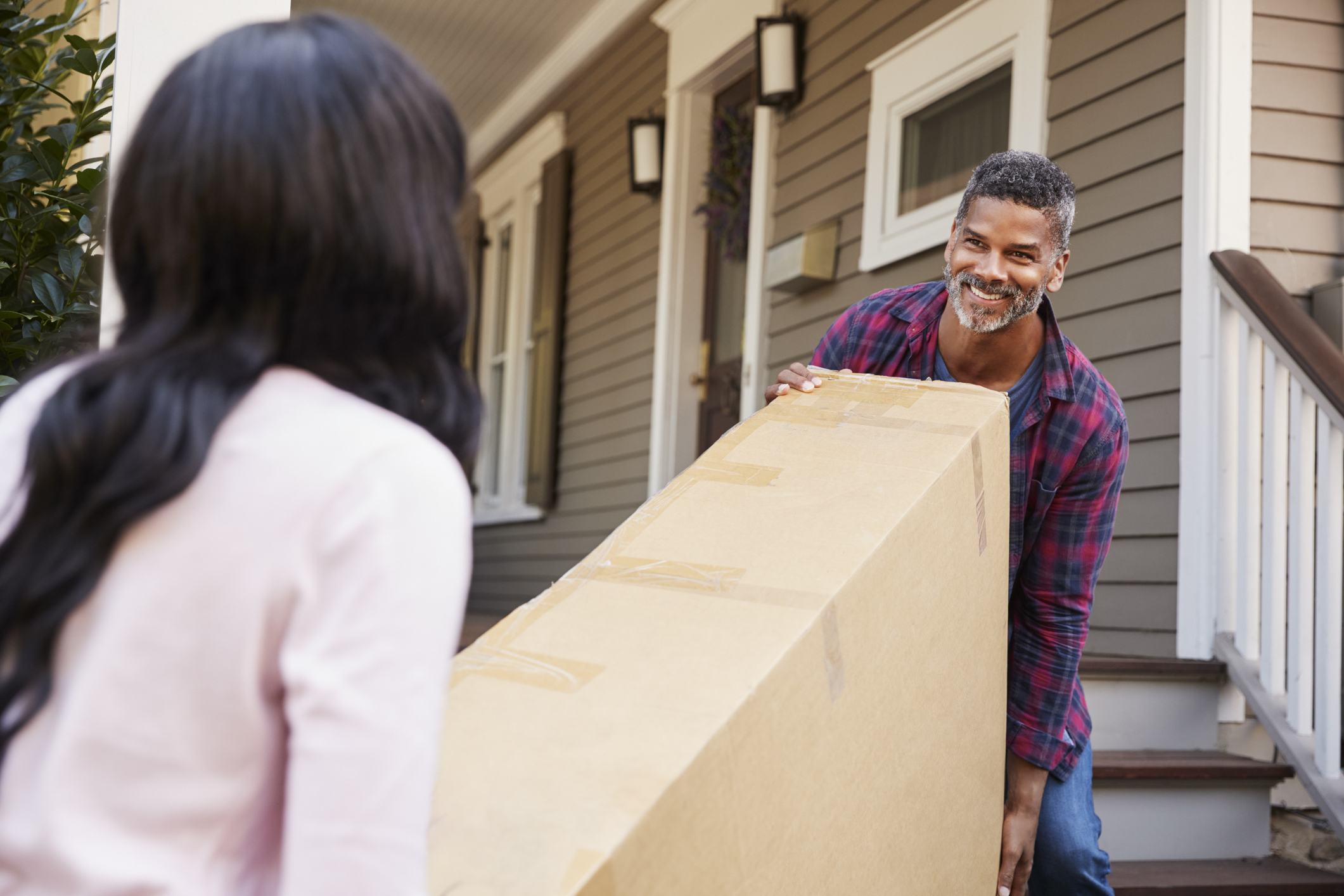 A man and a woman smile while carrying a large box into a house.